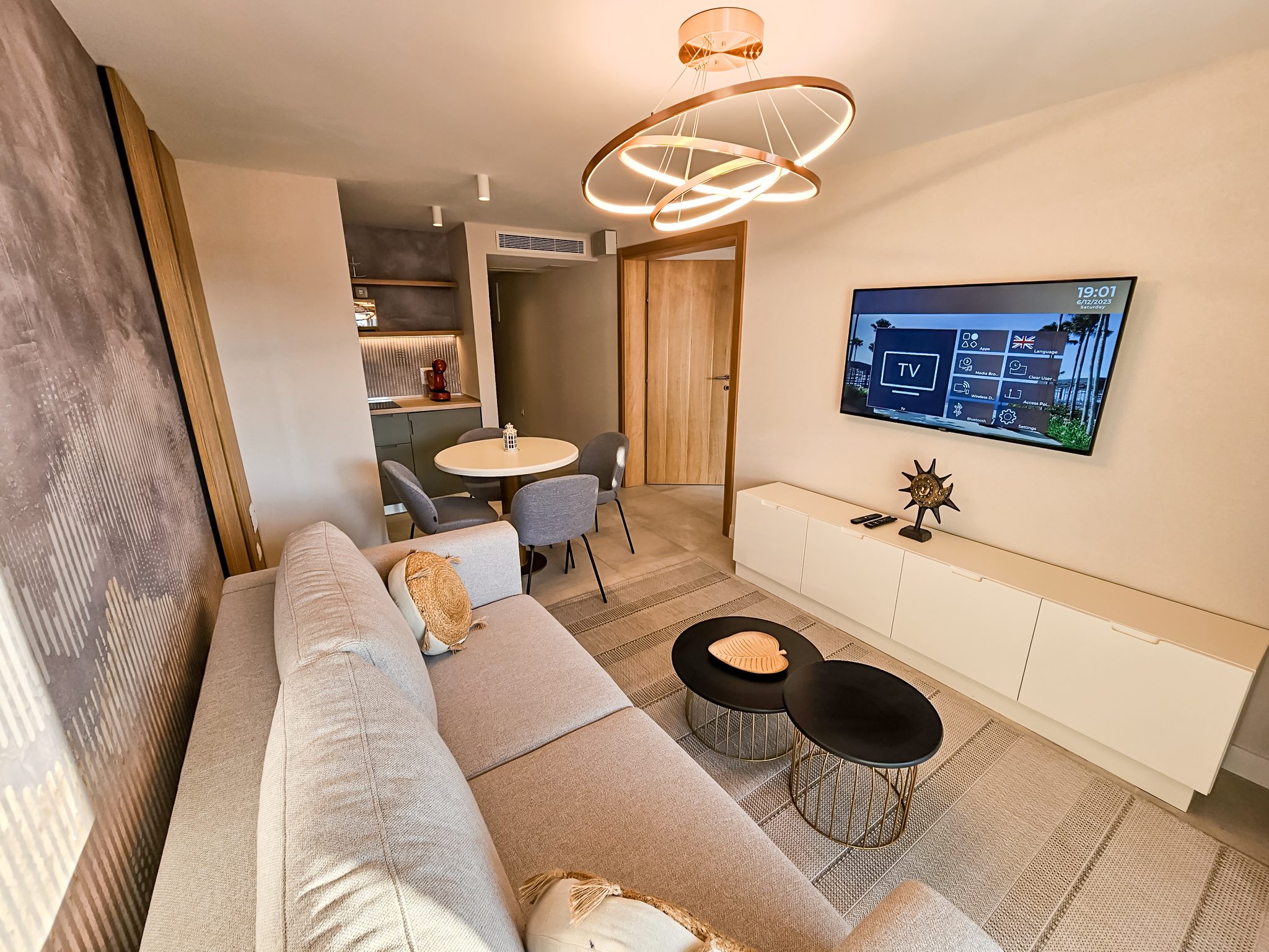 Moderna hotelska suite sa sivom sofom, kuhinjom i blagavaonom, osvetljena zlatnom lustre