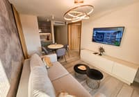 Moderna hotelska suite sa sivom sofom, kuhinjom i blagavaonom, osvetljena zlatnom lustre